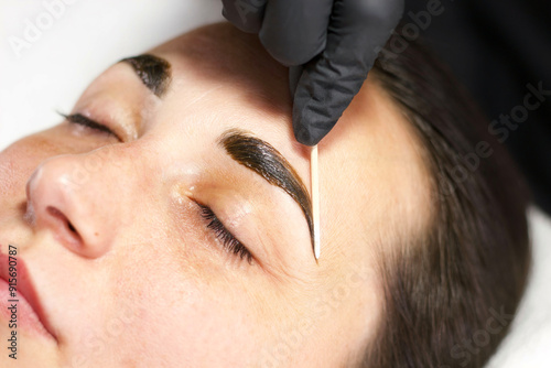  A close-up shot of a beauty professional using a cotton swab to apply tint to a woman's eyebrows. The woman’s eyes are closed, highlighting the meticulous attention to detail during the beauty proced