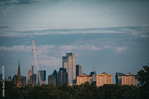Skyline of a modern city featuring high-rise buildings, construction cranes, and architectural landmarks, framed by trees under a serene, cloud-streaked sky.