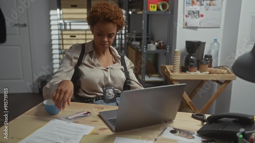 A focused african american female detective analyzes evidence at her office desk, indicating an investigation.