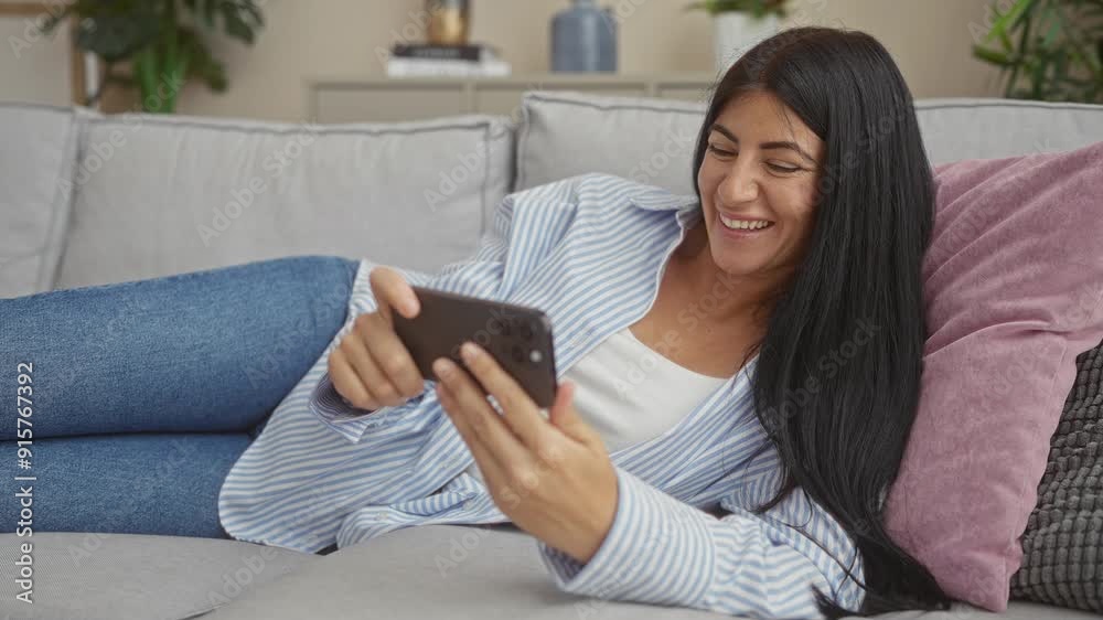 Young brunette hispanic woman lounging on couch in living room, smiling while using smartphone, suggesting a comfortable and relaxed home atmosphere. Young brunette hispanic woman lounging on couch in living room, smiling while using smartphone, suggesting a comfortable and relaxed home atmosphere.
