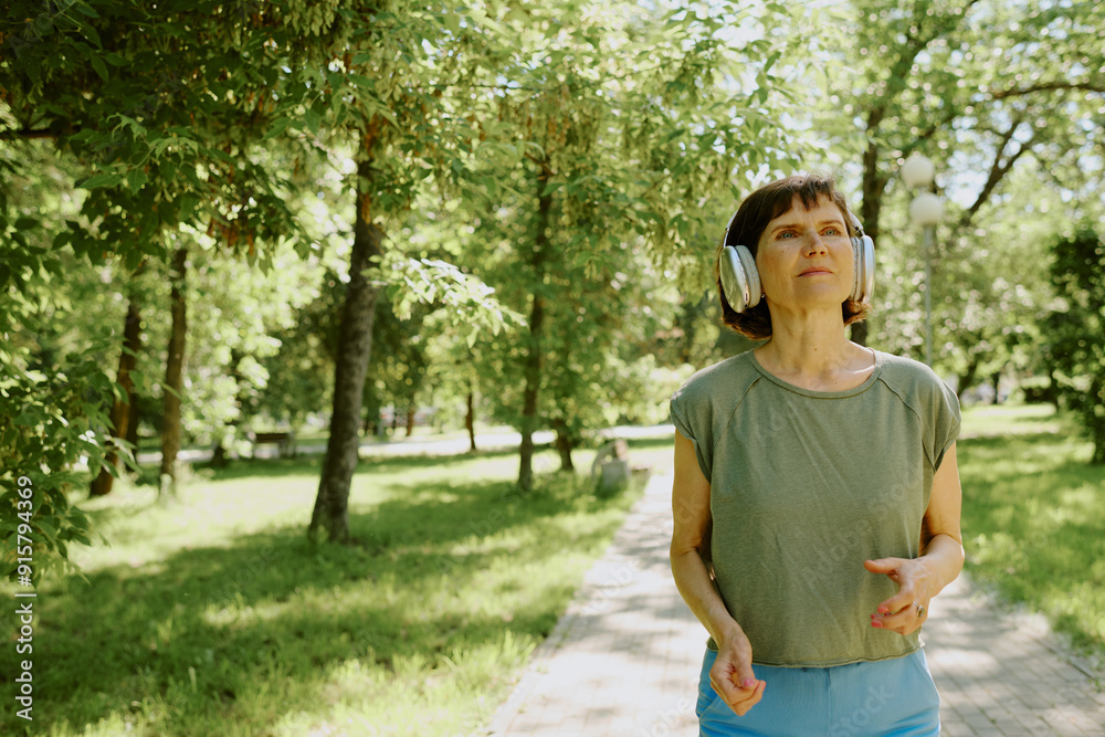 Senior brunette woman wearing silver headphones and doing cardio in park Senior brunette woman wearing silver headphones and doing cardio in park