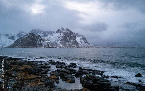 A scenic coastal view featuring a snow-capped mountain, rocky beach, stormy sky, and dynamic ocean, Lofoten, Norway