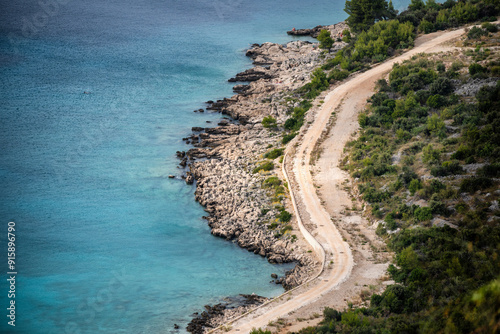 Fototapeta Naklejka Na Ścianę i Meble -  Bending, gravel road passing next to the beautiful, turquoise sea and wild beach at the small town of Primosten, Croatia