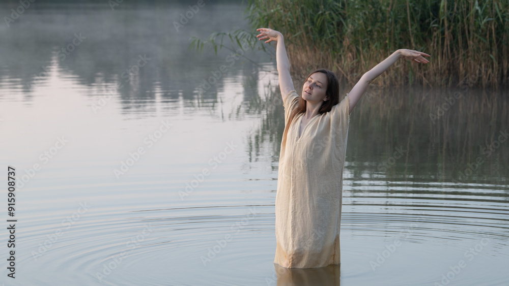 Fototapeta premium A dreamy girl or young woman with open arms in a simple light dress walking or dancing in a lake amidst the mist