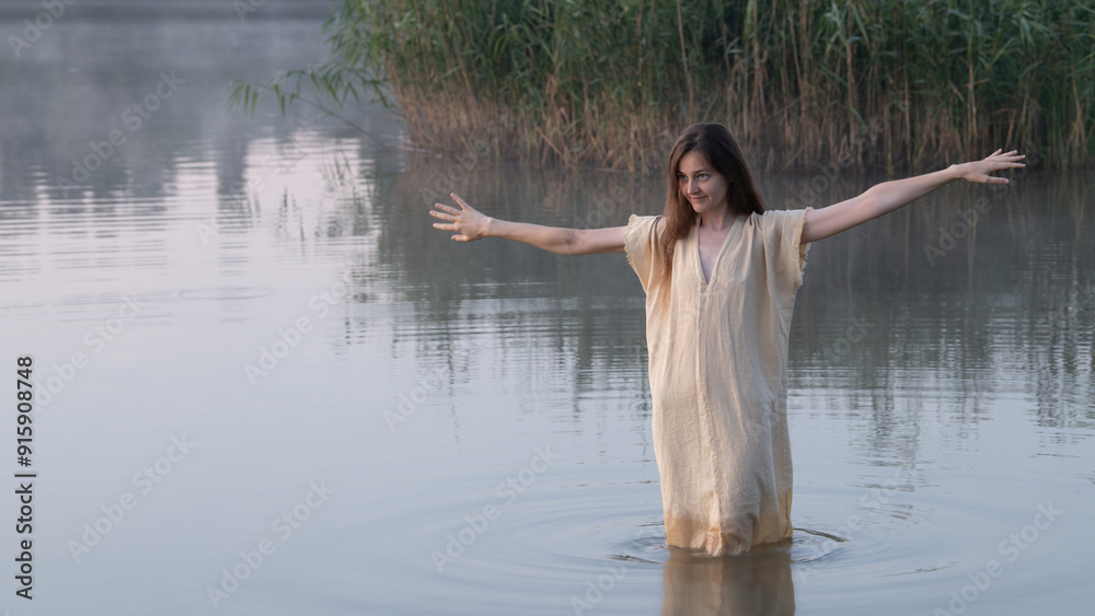 A dreamy girl or young woman with open arms in a simple light dress walking or dancing in a lake amidst the mist A dreamy girl or young woman with open arms in a simple light dress walking or dancing in a lake amidst the mist