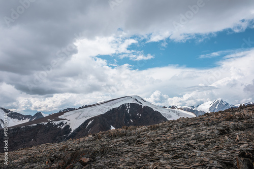 Sharp stones on high stony mountain pass with view to big glacier tongue among rockies and large snow-capped pinnacle in cloudy sky. Dramatic landscape in high rocky snow mountains in rainy weather. photo