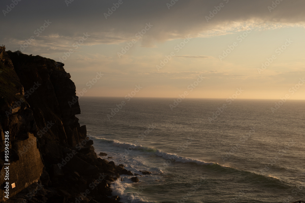 Golden hour paints the sky as a rugged cliff stands guard over the crashing waves Golden hour paints the sky as a rugged cliff stands guard over the crashing waves
