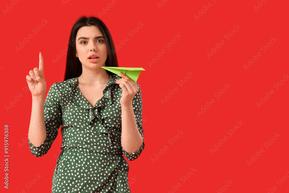 Shocked young woman with paper plane pointing at something on red background Shocked young woman with paper plane pointing at something on red background