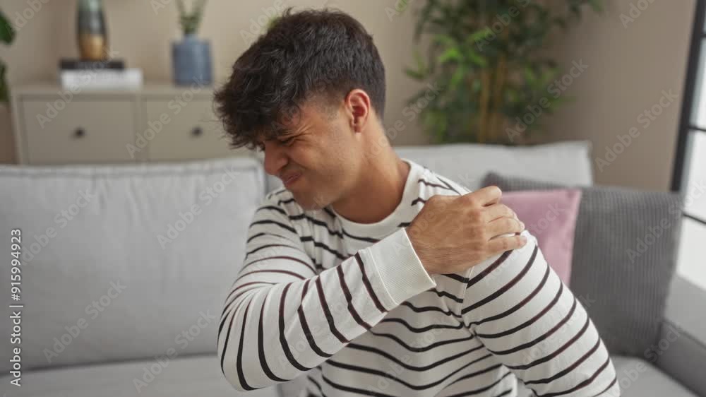 A young hispanic man in a striped shirt holds his shoulder in pain, sitting on a grey couch in a modern, well-decorated living room. A young hispanic man in a striped shirt holds his shoulder in pain, sitting on a grey couch in a modern, well-decorated living room.