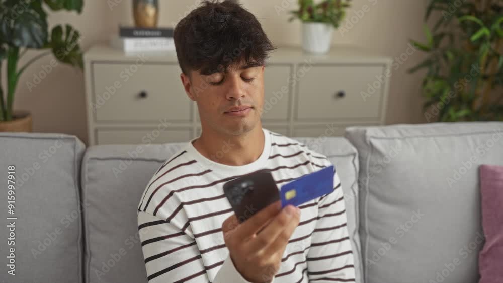 A young hispanic man in a striped shirt holds a credit card and phone while sitting on a gray couch in his home living room with modern decor and plants around. A young hispanic man in a striped shirt holds a credit card and phone while sitting on a gray couch in his home living room with modern decor and plants around.