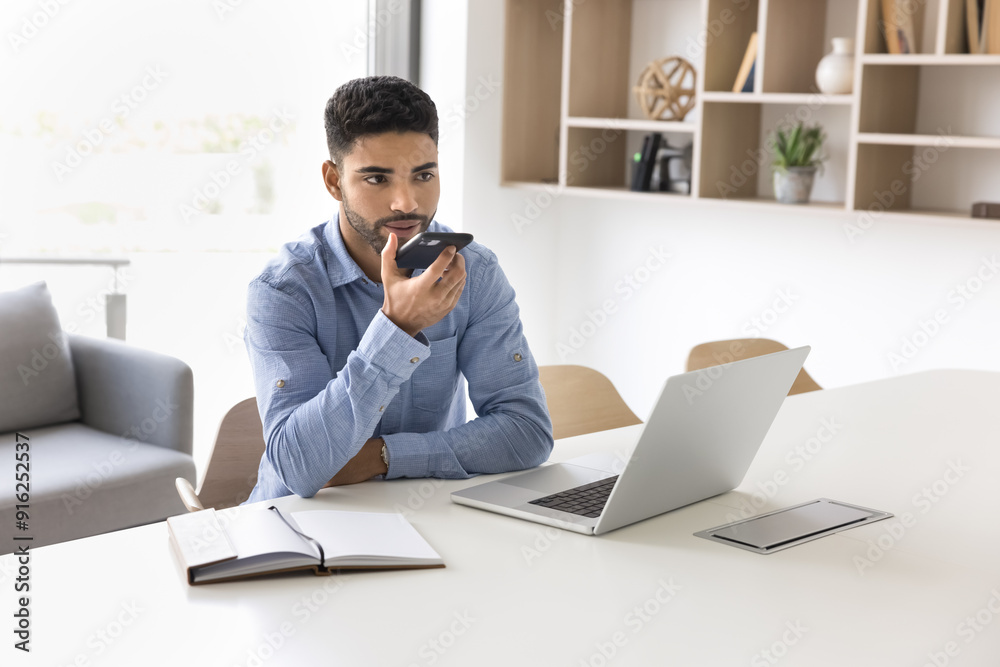 Serious young Arab businessman in casual speaking at mobile phone at office workplace table with laptop, recording voice message, using gadget for online work communication Serious young Arab businessman in casual speaking at mobile phone at office workplace table with laptop, recording voice message, using gadget for online work communication