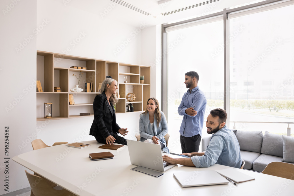 Happy diverse business team meeting in white colored modern office interior, sitting, standing at large table, talking, laughing, having fun, brainstorming on project, talking, discussing ideas