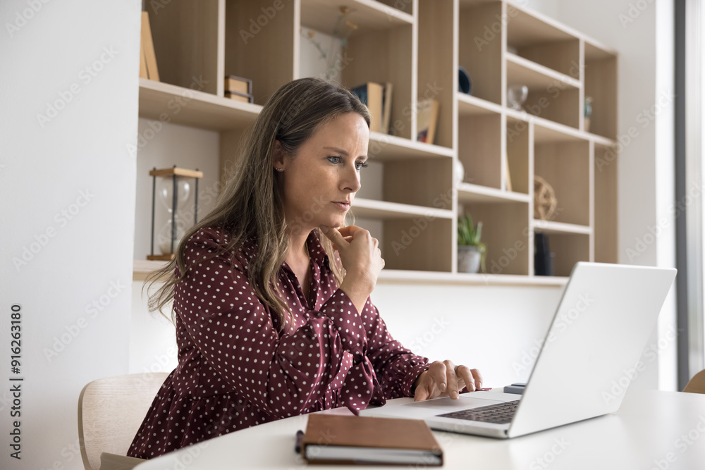 Focused mid adult business professional woman in casual working at laptop at workplace table, touching chin, thinking on career, modern technology for job, reading online content Focused mid adult business professional woman in casual working at laptop at workplace table, touching chin, thinking on career, modern technology for job, reading online content