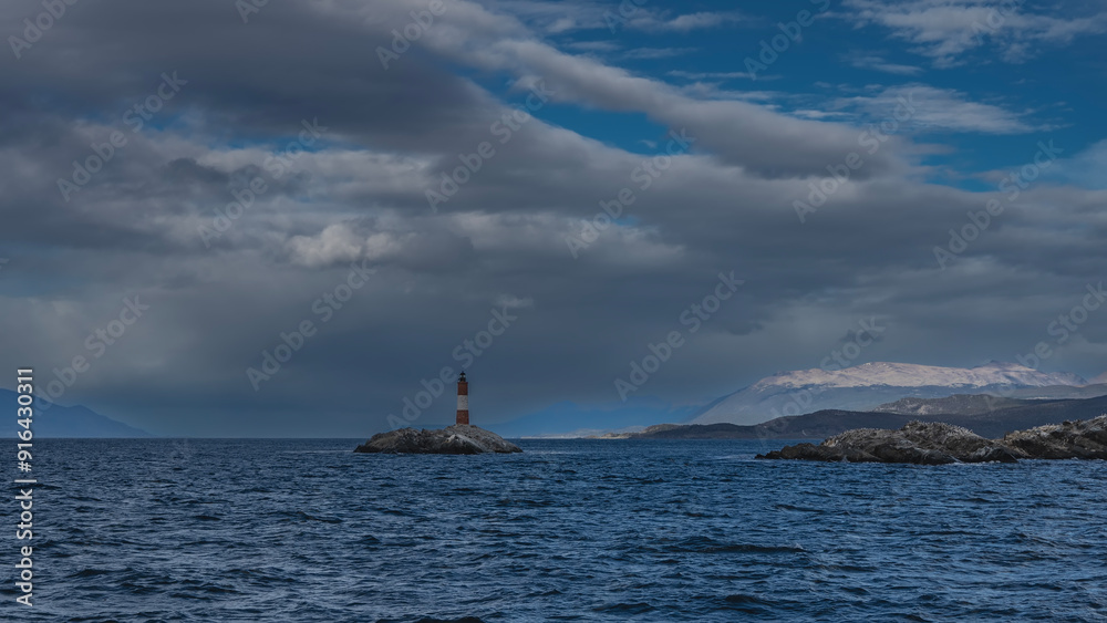 An old lighthouse on a tiny rocky island in the ocean. A red and white striped tower against the sky, clouds. Mountains in the distance. Ripples on the surface of the blue water of the Beagle Channel An old lighthouse on a tiny rocky island in the ocean. A red and white striped tower against the sky, clouds. Mountains in the distance. Ripples on the surface of the blue water of the Beagle Channel