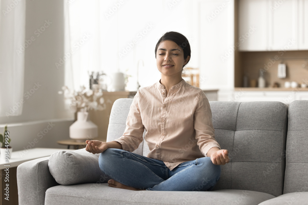 Peaceful young Indian woman sitting on cozy sofa in lotus pose, enjoy meditation with eyes closed, lead healthy lifestyle, relax, doing yoga for mental balance, relieves stress and negative emotions Peaceful young Indian woman sitting on cozy sofa in lotus pose, enjoy meditation with eyes closed, lead healthy lifestyle, relax, doing yoga for mental balance, relieves stress and negative emotions