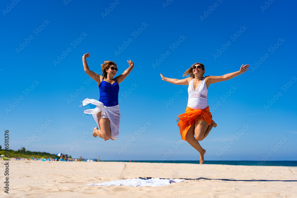Joyful summer. Two young women wearing swimsuits and sarong and sunglasses jumping on sandy beach against blue sky. Energy, happiness and activity. Joyful summer. Two young women wearing swimsuits and sarong and sunglasses jumping on sandy beach against blue sky. Energy, happiness and activity.