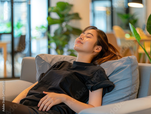 A young Asian woman is lying comfortably on a sofa with her eyes closed, enjoying a moment of relaxation.