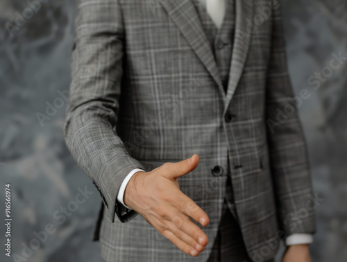 A man in a grey plaid suit extends his hand in a handshake gesture.