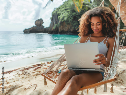 A woman with curly hair is smiling and working on her laptop while relaxing in a hammock on a tropical beach.