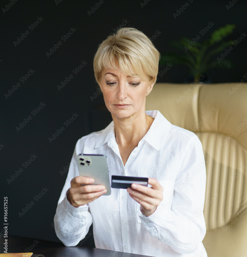 A woman carefully checks her phone while holding a credit card, engaged in an online transaction at her sleek office workspace. A woman carefully checks her phone while holding a credit card, engaged in an online transaction at her sleek office workspace.