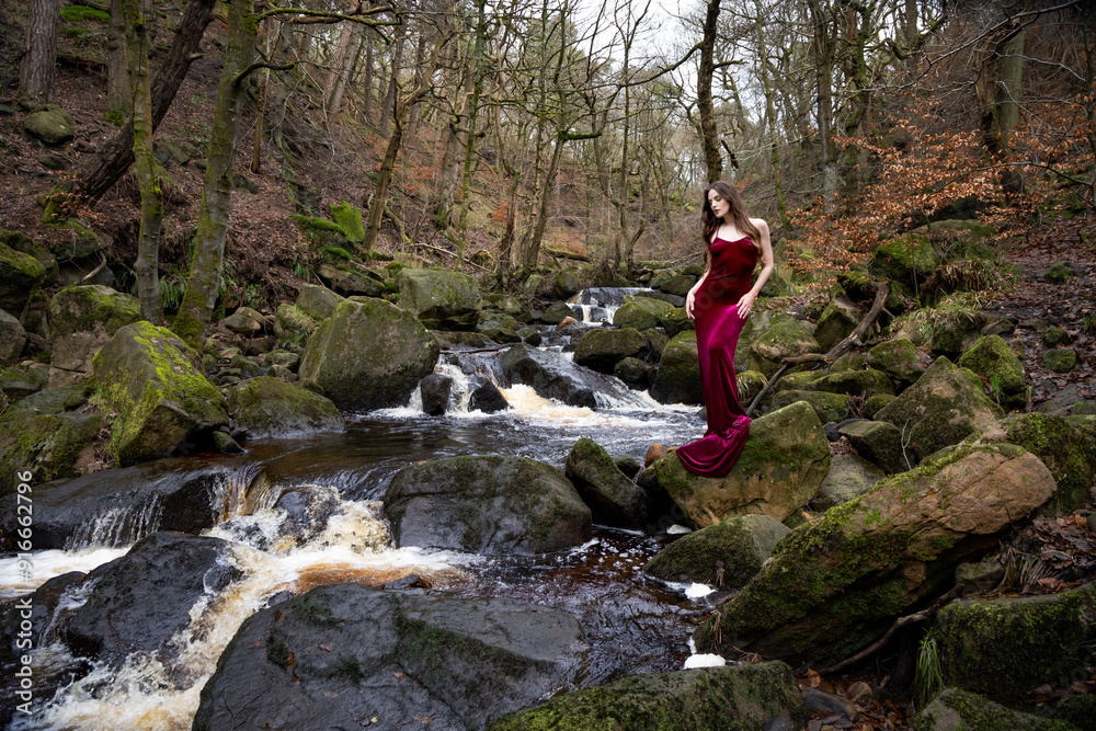 Fototapeta premium Beautiful girl with long dark hair, wearing a long red velvet dress, standing on a rock above a waterfall