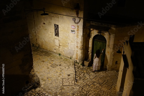Girl in white dress knocking on the door in a dark alley of Matera town in the night, Basilicata, Italy