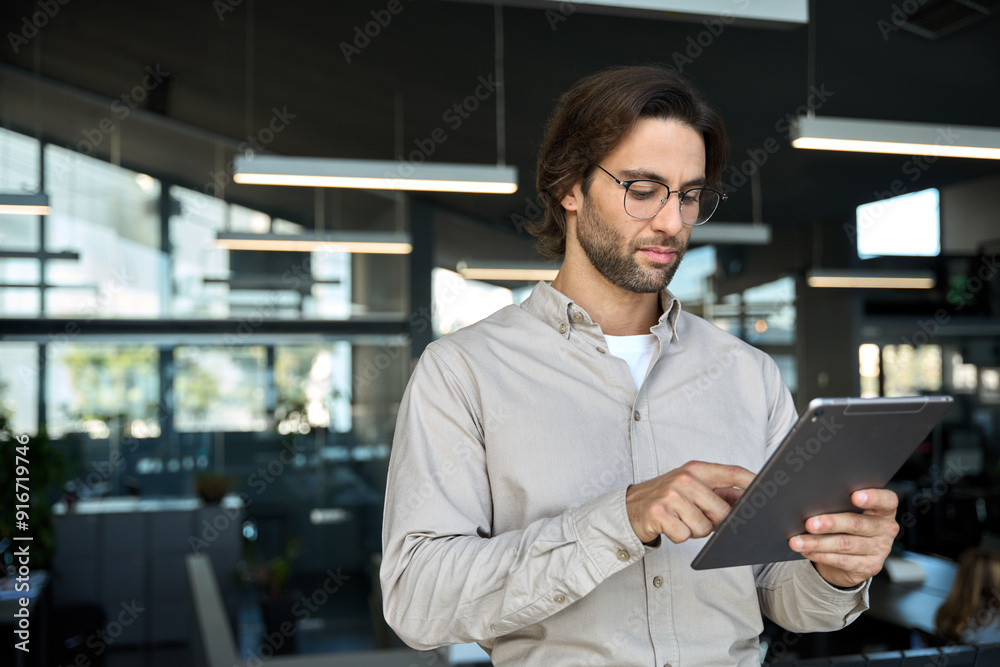Serious busy young European business man entrepreneur using tablet standing in office at work. Male professional executive manager using tab computer managing financial banking data. Copy space. Serious busy young European business man entrepreneur using tablet standing in office at work. Male professional executive manager using tab computer managing financial banking data. Copy space.