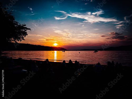 Ein atemberaubender Sonnenuntergang am See, mit Booten auf dem Wasser und einer dramatischen Wolkenlandschaft, die die friedliche Stimmung der Natur einfängt. photo