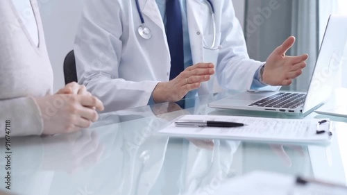 Doctor is consulting patient in modern fair clinic. Healthcare professional discusses medical information with a woman, gesturing above the laptop computer on the glass desk. Medicine photo