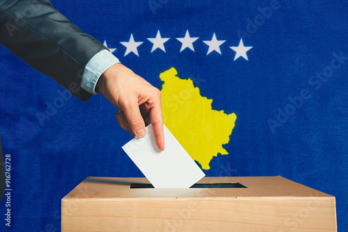 A hand holds a ballot in a ballot box against the background of the Kosovo flag. People place ballots in a ballot box with the Kosovo flag in the background.