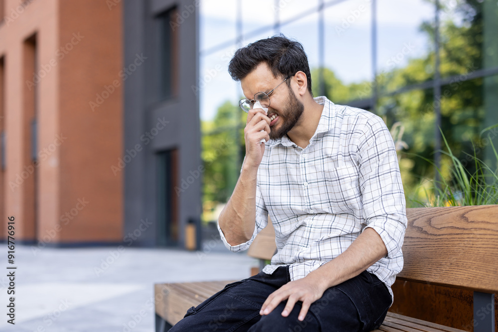 Man sneezing due to allergy while sitting outdoors on bench next to modern building Man sneezing due to allergy while sitting outdoors on bench next to modern building