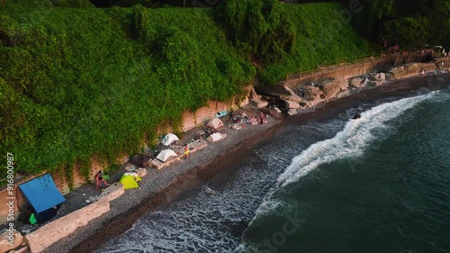 aerial shot of a tent camping on the seashore near a cliff covered with greenery drone shot of several tents standing on the seashore and waves. drone rises, camera looks down