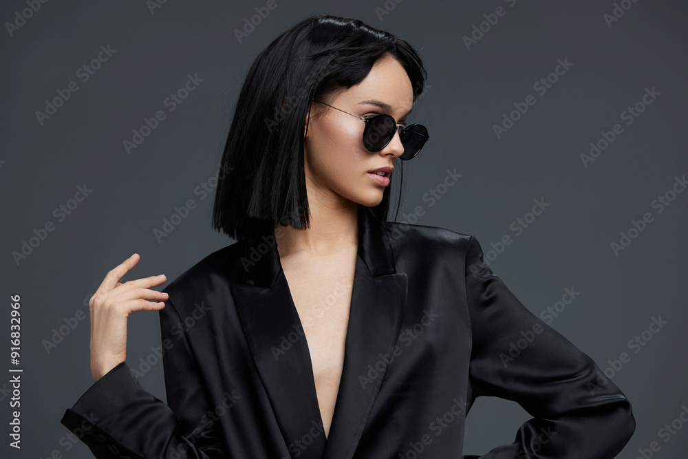 Asian woman in a black suit and sunglasses striking a pose against a gray background, exuding confidence and elegance