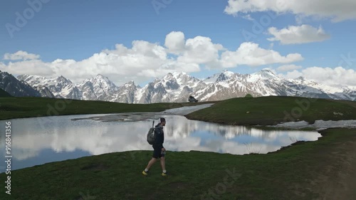 Aerial dronie of hiker with backpack. Person walking by Koruldi lakes in Svaneti mountains. Snow-capped peaks, blue sky, green landscape and tranquil water. Hiking eco tourism, adventure travel. Drone photo