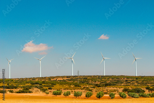 Photograph of industrial metal windmills lined up across the Castilian fields in Spain on a sunny day, with trees and plowed earth in the foreground, capturing a rural Spanish scene