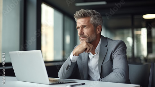 Portrait of a middle-aged business man sitting at a desk with a laptop in a modern office, looking away thinking about something. 