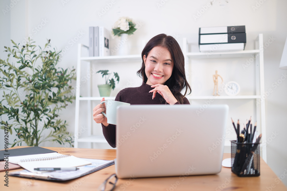 Young professional woman sits at her home office desk, smiling as she holds a coffee mug and looks at her laptop. Young professional woman sits at her home office desk, smiling as she holds a coffee mug and looks at her laptop.