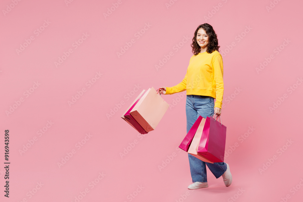Full body side view young woman wear yellow sweatshirt casual clothes hold in hand paper package bags after shopping isolated on plain light pastel pink background. Black Friday sale buy day concept. Full body side view young woman wear yellow sweatshirt casual clothes hold in hand paper package bags after shopping isolated on plain light pastel pink background. Black Friday sale buy day concept.