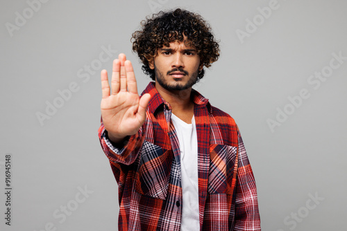 Young sad strict serious mad Indian man he wearing red shirt casual clothes showing stop gesture with palm looking camera isolated on plain grey color background studio portrait. Lifestyle concept.