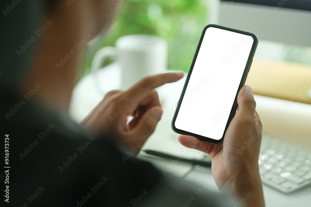 Man using smart phone while sitting front of laptop at office desk. Over shoulder view.