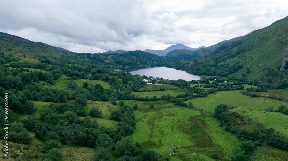Obraz premium A stunning aerial view of a lush green valley with a serene lake in the distance, surrounded by rolling hills and dense forests under a cloudy sky.