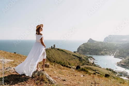 Happy woman in a white dress and hat stands on a rocky cliff above the sea, with the beautiful silhouette of hills in thick fog in the background.