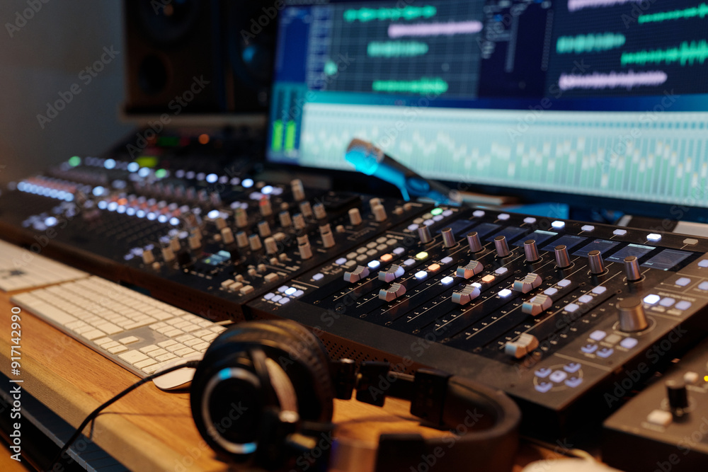 Professional audio mixing board with multiple channels illuminated, showing various sound frequencies on screen in background with headphones resting on desk nearby Professional audio mixing board with multiple channels illuminated, showing various sound frequencies on screen in background with headphones resting on desk nearby