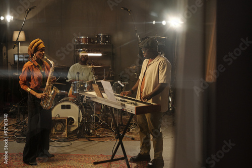 Musicians jamming in studio with saxophonist, drummer, and keyboardist engaged in passionate performance under warm lighting creating an intimate atmosphere