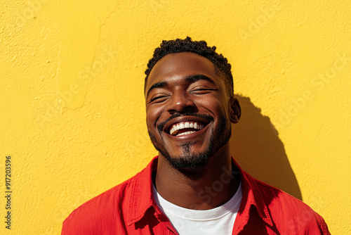 Black Man Expressing Joyful Laughter in a Close-Up Headshot, Sunlight Highlighting His Face, Red Shirt and White T-Shirt Against a Yellow Wall