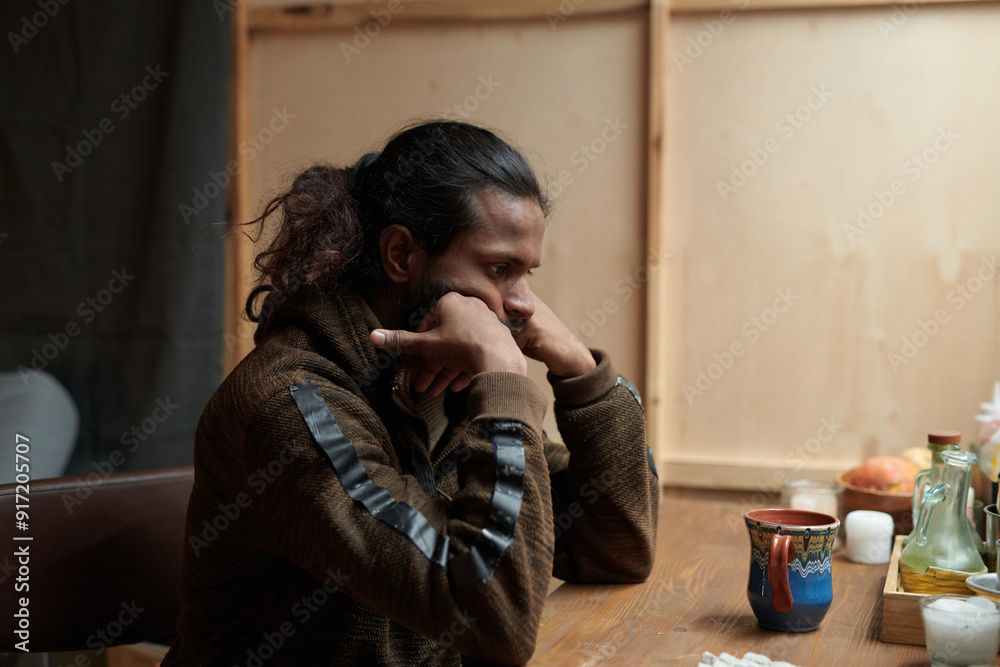 Pensive young man sitting at wooden table in rustic kitchen setting, resting chin on hand with thoughtful expression, warmly lit space with various condiments and jars visible Pensive young man sitting at wooden table in rustic kitchen setting, resting chin on hand with thoughtful expression, warmly lit space with various condiments and jars visible