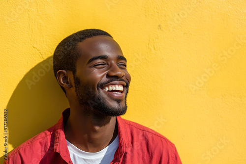 Cinematic Headshot of a Black Man with a Radiant Smile, Sunlit Face, Wearing a Red Shirt Over a White T-Shirt, Set Against a Yellow Wall 