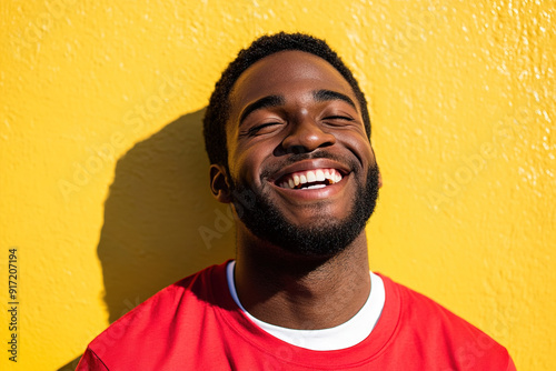 Cinematic Headshot of a Black Man with a Radiant Smile, Sunlit Face, Wearing a Red Shirt Over a White T-Shirt, Set Against a Yellow Wall 