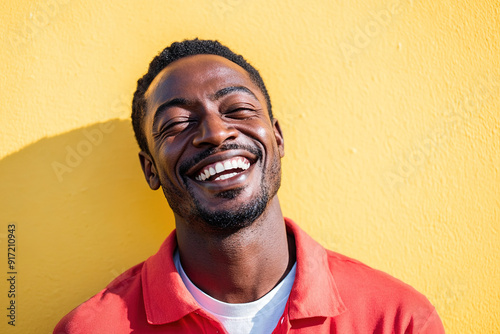 Joyful Black Man Laughing with Sunlight on His Face, Wearing a Red Shirt and White T-Shirt, Headshot Against a Bright Yellow Wall Background