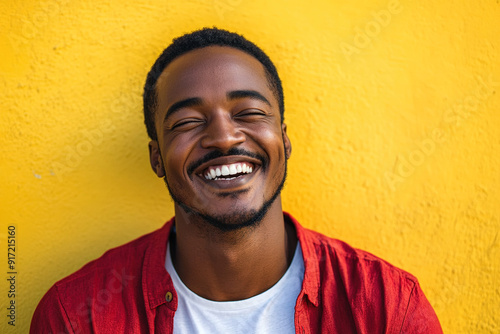 Smiling Black Man in Red Shirt with White T-Shirt Underneath, Laughing Joyfully on a Sunny Day with Sunlight Illuminating His Face, Against a Yellow Wall Background. High-Resolution Headshot Portrait 